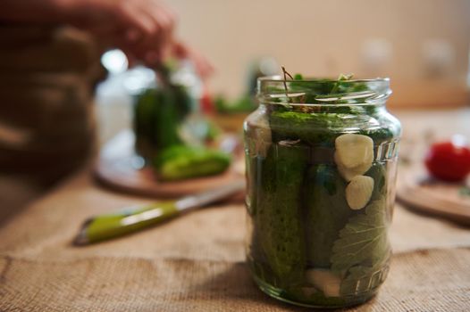 Focus on a glass jar, filled with freshly pickled cucumbers, herbs and fresh garlic cloves, on a blurred background of a woman, housewife making pickles at home kitchen. Marinating, canning, pickling.