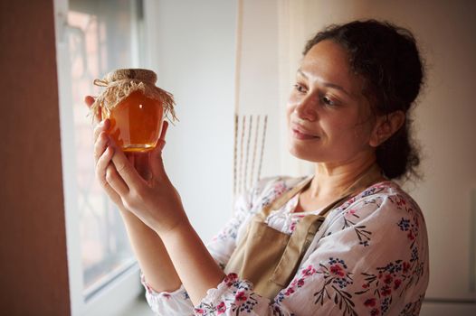 Focus on freshly cooked and canned homemade peach jam in a glass jar with a burlap on the lid, in the hands of a pleasant and happy multiethnic woman, standing by the window at home kitchen interior