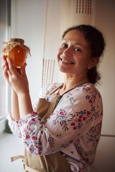 Beautiful multiethnic woman, a pleasant housewife, in a beige apron, holds a jar of homemade peach jam, canned according to a traditional family recipe, and smiles cutely, looking at the camera