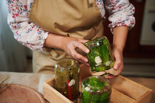 Details: Hands of a housewife turning over the glass jars of pickled cucumbers covered with pickling marinade, placing them with the lid down on wooden crate in the home kitchen. Harvest conservation.