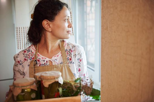 Pleasant multiethnic women, housewife in chef's apron thoughtfully looks through the window in the kitchen while holding a box with jars of pickled vegetables, preserved according to a homemade recipe
