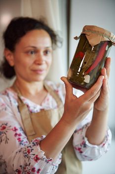 Focus on a glass jar of homemade pickled red and green chili peppers, marinated according a traditional family recipe in the hands of a multiethnic cute housewife, standing by a window at home kitchen