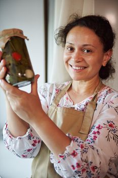 Portrait of happy multiethnic cute woman, housewife showing jar of pickled chili peppers, marinated with brine, umbrella dill, garlic and fragrant culinary herbs, according a traditional family recipe
