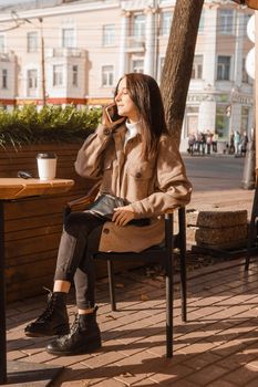 A stylish brunette woman is sitting at a table in an outdoor cafe with coffee and a phone. The girl is talking on the phone. Autumn walk.