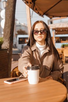 A stylish brunette woman in sunglasses is sitting at a table in an outdoor cafe with coffee and a phone. The girl is talking on the phone. Autumn walk.