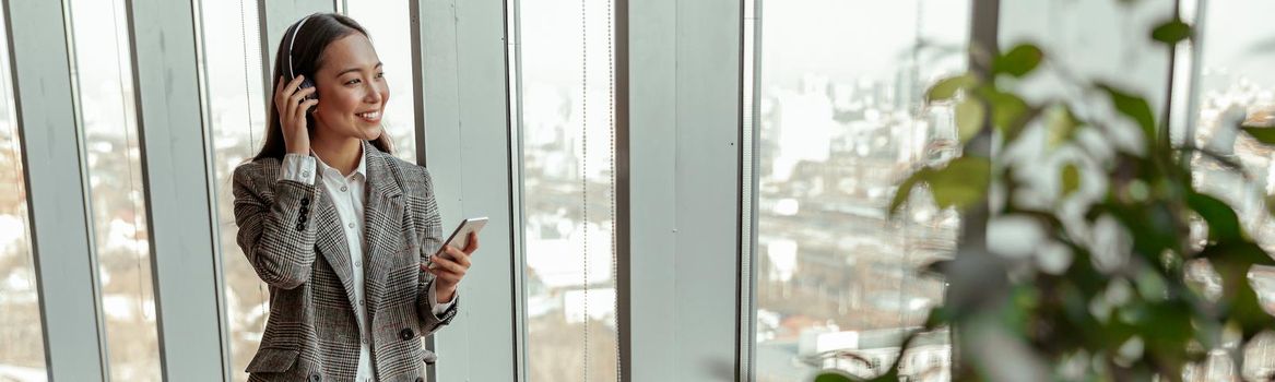 Smiling Asian Business woman listening music from phone in headphones standing near window in office