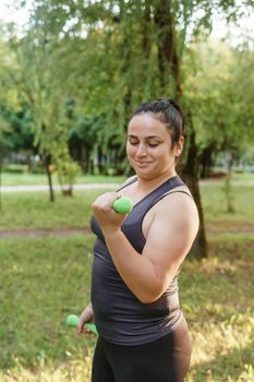 A charming brunette woman plus-size body positive practices sports in nature. Exercises with dumbbells.