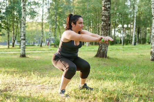 A charming brunette woman plus-size body positive practices sports in nature. Exercises with dumbbells.