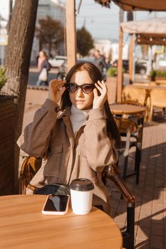 A stylish brunette woman in sunglasses is sitting at a table in an outdoor cafe with coffee and a phone. The girl is talking on the phone. Autumn walk.