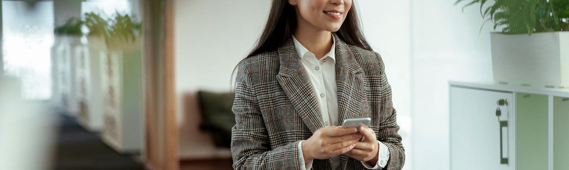 Portrait of attractive asian business woman with phone standing in modern office and looking away