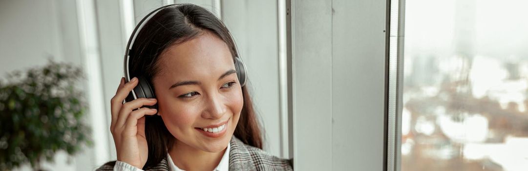 Asian Business woman listening music from phone in headphones standing near window in office