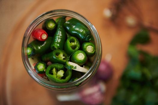 Selective focus. Jar of freshly marinated chili peppers, poured with boiling marinade or brine, on a wooden chopping board with blurred d culinary herbs and garlic cloves. Top view. Still life. Food