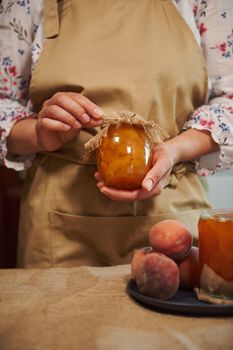 Details: Hands of a woman, housewife, pastry chef, confectioner in beige apron, holding jar of homemade jam from juicy ripe organic peaches, canned according to traditional family recipe. Canned food