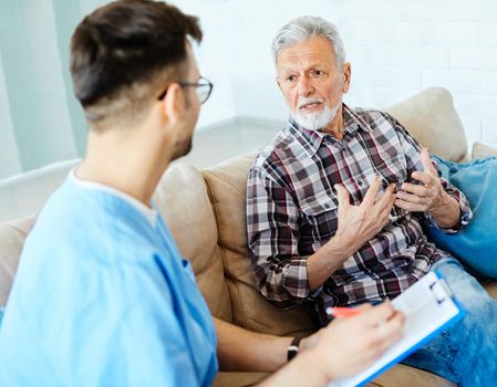 Doctor or nurse caregiver talking with senior man with chest pain and coughing problems on sofa at home or nursing home