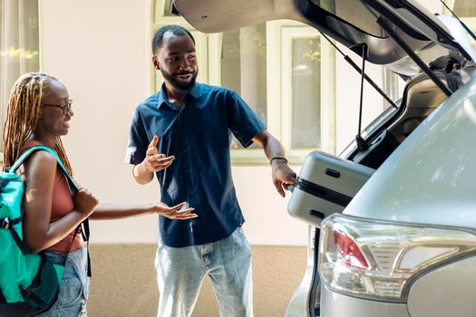 Happy people loading baggage in car trunk, travelling on summer vacation with trolley and bags. Couple leaving on holiday adventure with vehicle, feeling excited about destination.