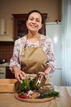 Charming multi-ethnic woman, housewife stands in the home kitchen interior and smiles looking at camera, showing jars with homemade pickled cucumbers and chili peppers. Seasonal canning of vegetables