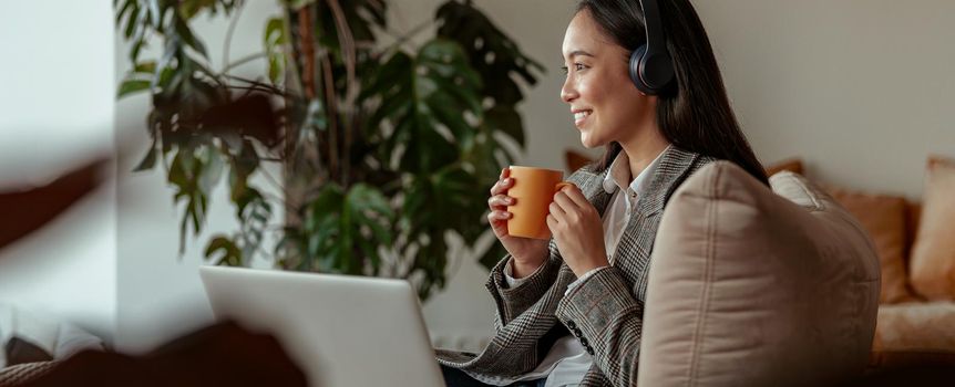 Asian business woman listening music on headphones sitting at laptop with coffee in cozy office