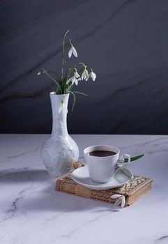Still life with white snowdrops in a ceramic vase in the form of a swan, coffee in a white cup, conceptual morning ritual whith a book.High quality photo