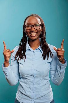 Excited happy beautiful woman smiling heartily while pointing fingers up on blue background. Confident and positive young adult person pointing hands up while looking at camera.