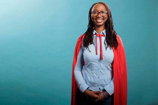 Positive smiling heartily mighty and brave justice defender standing on blue background while looking at camera. Young adult confident and strong superhero woman wearing hero cloak. Studio shot