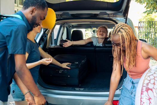 Multiethnic people travelling on summer holiday, loading baggage and inflatable in car trunk to travel to seaside. People preparing to leave on vacation trip with luggage in automobile.