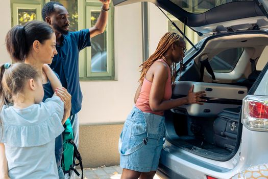 Diverse family and friends leaving on journey, loading inflatable and trolley bags in trunk of vehicle to travel on summer holiday vacation. Going to seaside destination with smiling people.