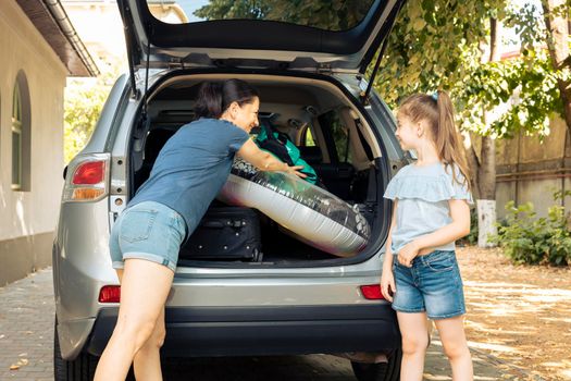 Mother and small kid loading baggage in automobile trunk to leave on holiday vacation at seaside. Travelling during summer with vehicle and luggage, suitcase, inflatable and trolley.
