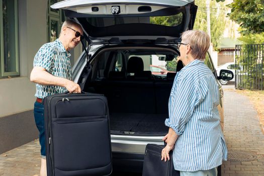 Retired man and woman loading luggage in car trunk, preparing to leave on road trip holiday. Travelling together during summer, going on vacation journey with suitcase, baggage and bags.
