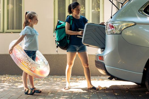 Mother and small kid travelling on vacation, loading baggage trolley and inflatable in vehicle trunk to go to seaside destination during summer holiday. Woman and little girl leaving on road trip.