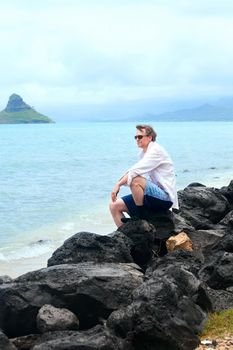 Handsome Caucasian man in forties sitting on rocky shore of hawaiian beach, by Chinaman's Hat island