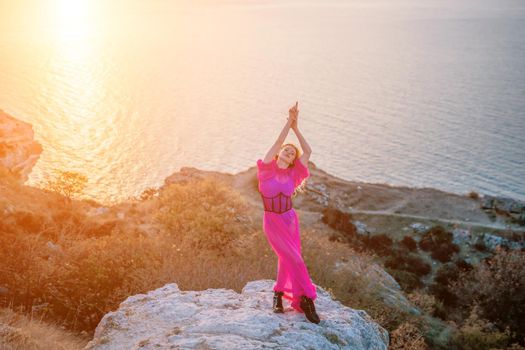 Side view a Young beautiful sensual woman in a red long dress posing on a volcanic rock high above the sea during sunset. Girl on the nature on blue sky background. Fashion photo