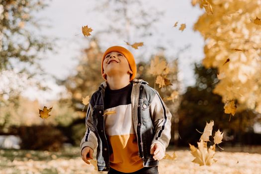 Kid having fun in autumn park with fallen leaves, throwing up leaf. Child boy outdoors playing with maple leaves