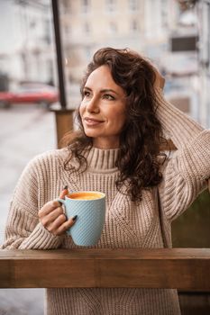 A middle-aged woman in a beige sweater with a blue mug in her hands is in a street cafe on the veranda.