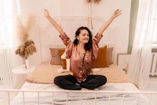 Photo of a middle aged woman in pajamas stretching her arms and smiling while sitting on the bed after sleeping or napping.