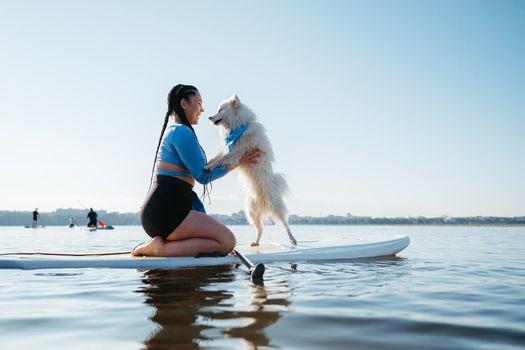 Happy Young Woman with Locs Hugging with Her Dog Japanese Spitz While Sitting on the Sup Board on Lake