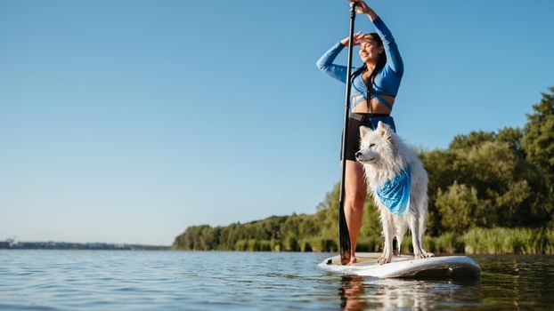 Happy Young Woman Enjoying Life on the Lake at Early Morning Standing on Sup Board with Her Dog Snow-White Japanese Spitz