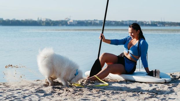 Young Woman Sitting on Sup Board and Laughing at Her Japanese Spitz Digging a Hole on Beach of City Lake, Paddleboarding with Dog