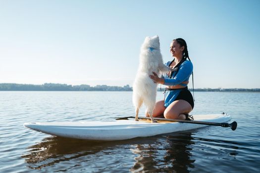 Happy Young Woman Hugging with Her Dog Japanese Spitz While Sitting on Sup Board on the City Lake at Sunrise