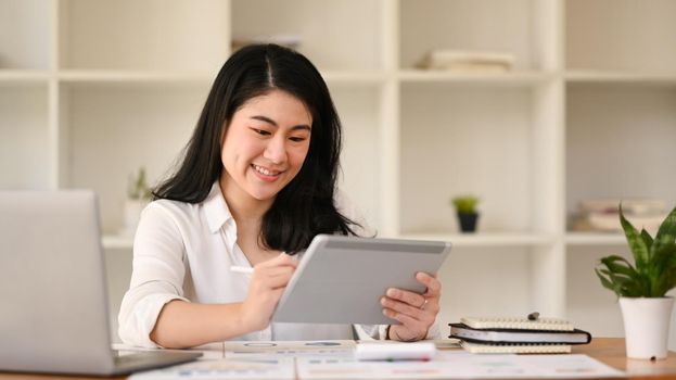 Cheerful asian female accountant checking online information on digital tablet, working at her workplace.