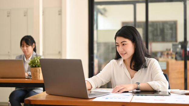 Asian female accountant using laptop and working with financial reports at her office desk.