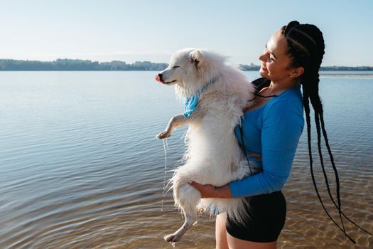 Happy Woman with Dreadlocks Holding on Hands Her Snow-White Dog Japanese Spitz on Lake at Sunrise