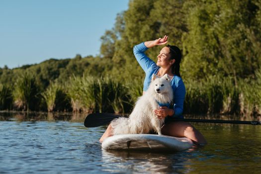 Happy Young Woman on the Lake at Early Morning Sitting on Sup Board with Her Dog Snow-White Japanese Spitz and Enjoying Sunrise