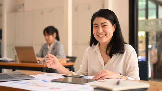 Happy asian female office worker sitting in corporate office ad smiling to camera.