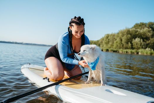 Cheerful Woman with Locs Helping Her Dog Japanese Spitz Get Out of the Water, While Sitting on Sup Board on the Lake