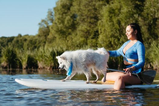 Young Woman Paddleboarding with Her Pet on the Lake, Snow-White Dog Breed Japanese Spitz Standing on Sup Board