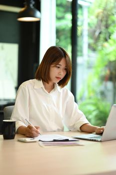 Concentrated female economist using laptop an working with document at office desk.