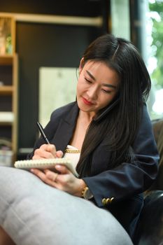 Asian working woman having phone conversation and making notes on notebook.