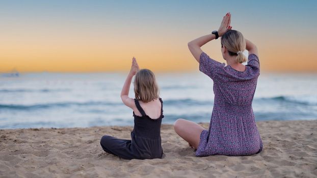 Mother and daughter do yoga on beach in evening. People have fun outdoors. Concept of summer holidays and friendly family and relaxation.