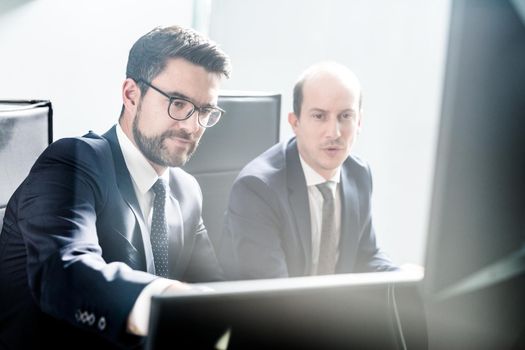 Image of two thoughtful businessmen looking at data on multiple computer screens, solving business issue at business meeting in modern corporate office. Business success concept.