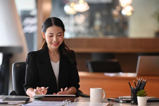 Cheerful businesswoman sitting in modern workplace and using digital tale.